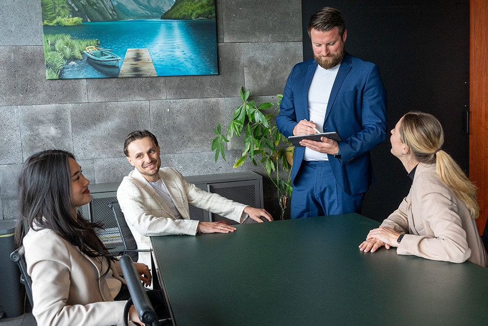 Ein Beratungsgespräch bei der André Lautermann Immobilienberatung. Ein Berater im blauen Anzug steht an einem dunkelgrünen Konferenztisch und macht sich mit einem Stift Notizen auf einem Tablet in seiner Hand. Drei weitere Personen – zwei Frauen und ein Mann in heller Kleidung – sitzen entspannt am Tisch und blicken aufmerksam und lächelnd zu ihm auf. Im Hintergrund der modernen Büroumgebung sind eine graue Steinwand, eine Pflanze und ein Landschaftsbild zu sehen.