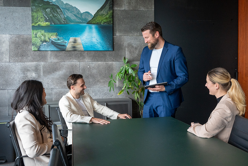 Ein freundliches Beratungsgespräch bei der André Lautermann Immobilienberatung. Ein Berater im blauen Anzug steht mit einem Tablet in der Hand an einem dunkelgrünen Konferenztisch und spricht lächelnd mit drei sitzenden Personen. Zwei Frauen und ein Mann in heller Kleidung blicken ihm aufmerksam und gut gelaunt zu. Die moderne Büroumgebung zeigt im Hintergrund eine graue Steinwand, eine Zimmerpflanze und ein Landschaftsbild.