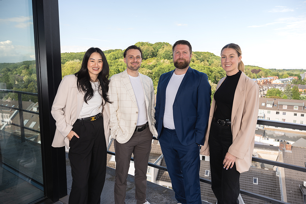 Ein Foto von vier professionell gekleideten Personen auf einer Dachterrasse. Von links nach rechts: Eine Asiatin mit langen dunklen Haaren in einem rosa Blazer und schwarzer Hose mit goldenem Gürtel. Ein Mann in einem hellen karierten Sakko und brauner Hose. Ein Mann mit Bart in einem dunkelblauen Anzug. Eine blonde Frau mit Pferdeschwanz in einem beigen Blazer und schwarzer Hose. Sie stehen auf einem Balkon mit Glas- und Metallgeländer und blicken über eine Stadt auf einen bewaldeten Hügel unter klarem, blauem Himmel. Alle vier lächeln freundlich in die Kamera.