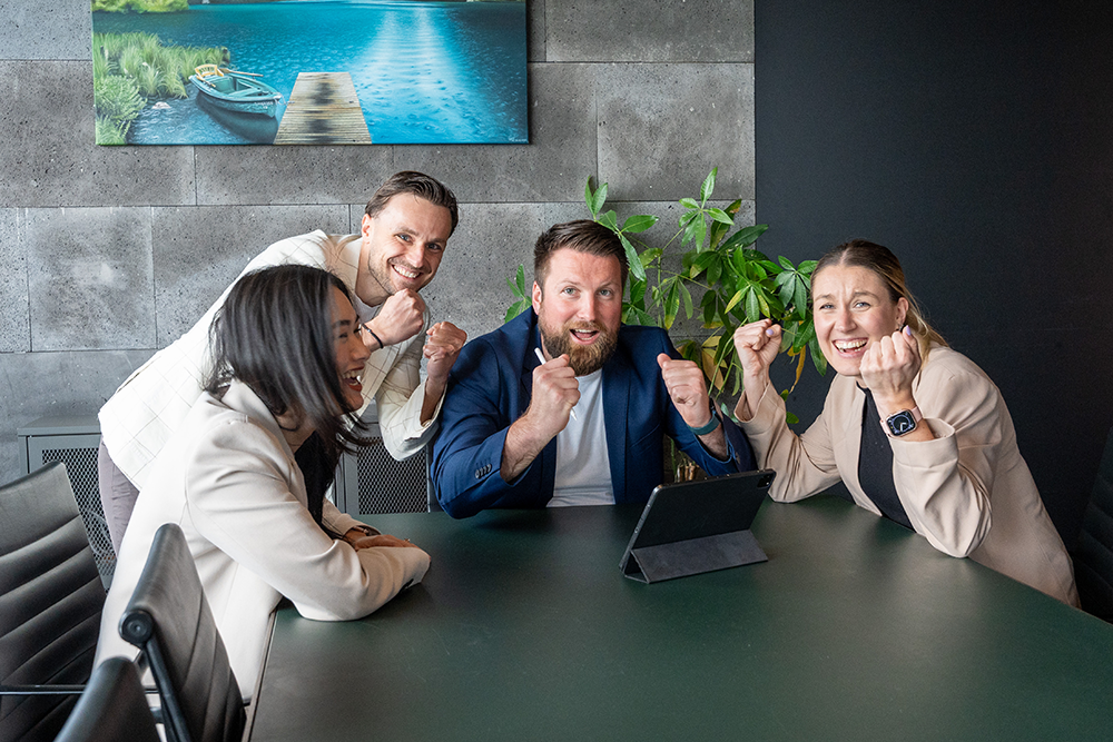 Ein Team-Foto von André Lautermann Immobilienberatung in einem modernen Konferenzraum, das einen gemeinsamen Erfolg feiert. Vier Teammitglieder (zwei Frauen und zwei Männer) umringen einen dunkelgrünen Tisch mit einem aufgestellten Tablet. Der Mann im blauen Anzug, der einen Stift hält, ballt triumphierend beide Fäuste, ebenso wie der Mann im hellen Sakko und die Frau in Beige. Alle lächeln breit und sehen begeistert und erfolgreich aus. Im Hintergrund sind eine graue Steinwand, eine große Pflanze und ein Landschaftsbild zu sehen.