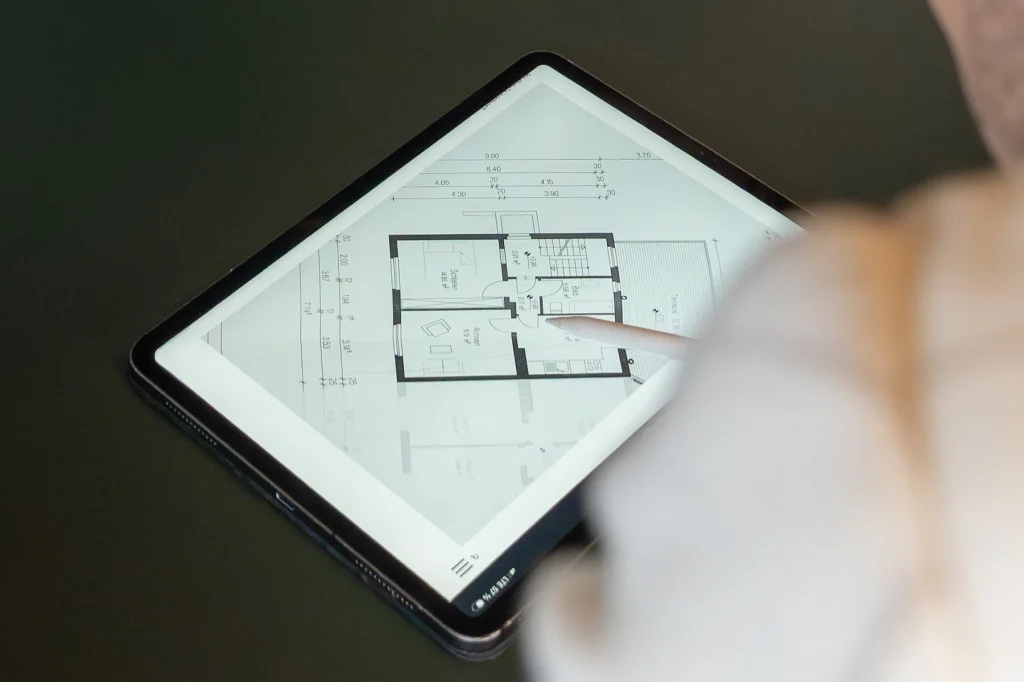 Eine Nahaufnahme aus der Vogelperspektive zeigt eine Person (teilweise sichtbar, mit einem Eingabestift in der Hand), die ein iPad Pro verwendet, das flach auf einem dunklen Schreibtisch liegt. Der Bildschirm des iPads zeigt einen detaillierten architektonischen Grundriss einer Wohnung. Der Eingabestift zeigt präzise auf den zentralen Wohnbereich des Grundrisses, was eine Verbindung zwischen Raumdaten und Wert darstellt. Die Beleuchtung ist weich und fokussiert, und ein Paar moderne Lesebrillen liegen unscharf im Hintergrund.
